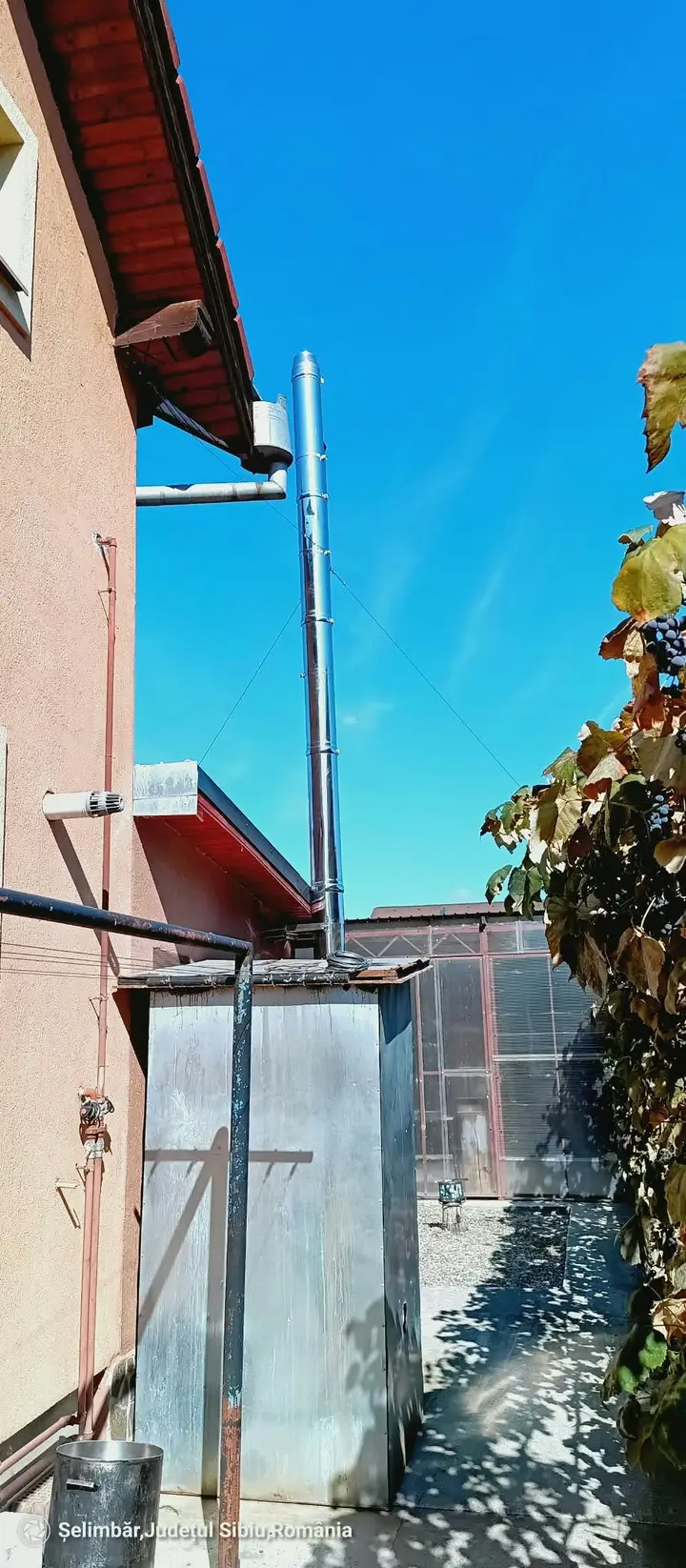 Coș din inox, lângă o anexă din tablă, cu o casă în fundal